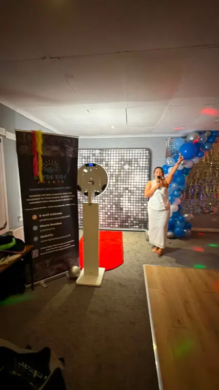 Halo selfie booth on a red carpet in front of a silver sequin shimmer wall, with a guest singing on the microphone beside a blue and silver balloon arch