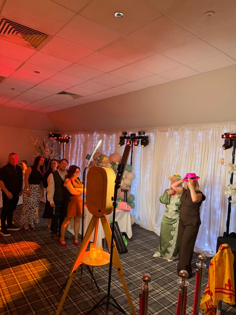 Vintage rustic wooden photo booth on a wooden tripod at a Scottish party, guests trying on colourful hats beside a white fairy-light drape and tartan carpet
