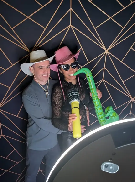 Wedding guests in cowboy hats posing with an inflatable saxophone and microphone at a Halo selfie booth against a navy and gold geometric backdrop