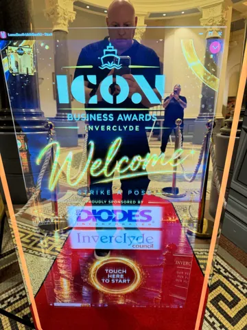 Magic Mirror photo booth with a personalised ICON Business Awards welcome screen behind a red carpet and gold rope stanchions in Greenock Town Hall