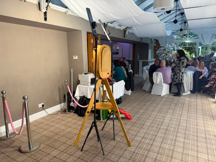 Vintage rustic wooden photo booth on a tripod with rope stanchions in a bright conservatory wedding reception as guests dine