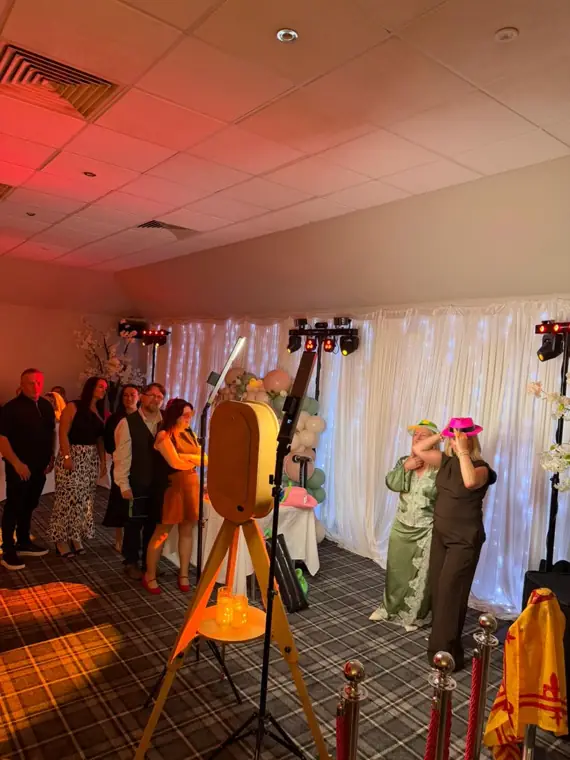 Vintage rustic wooden photo booth on a wooden tripod at a Scottish party, guests trying on colourful hats beside a white fairy-light drape and tartan carpet