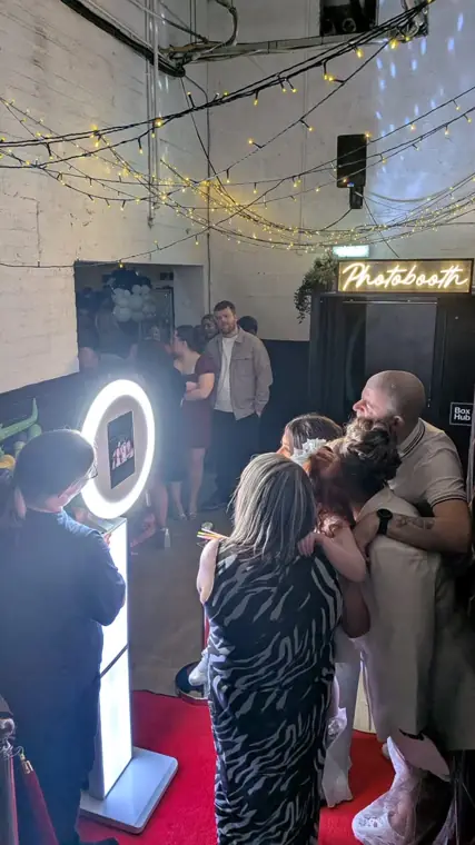 Halo selfie booth on a red carpet with rope stanchions under a neon Photobooth sign at a warehouse wedding, bride and guests hugging